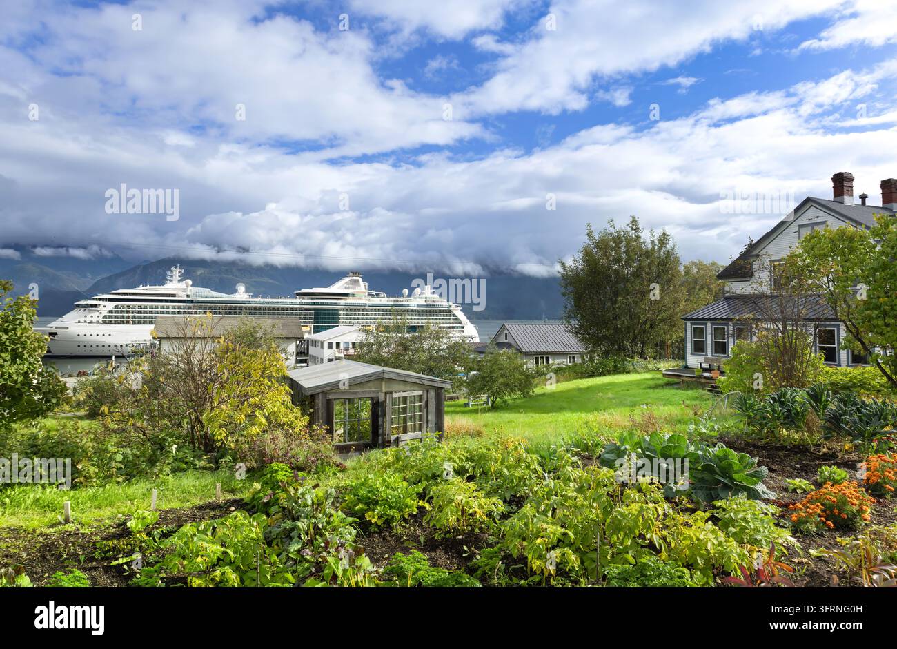Vista panoramica di Haines, Alaska, con una nave da crociera accanto al fiordo, case e giardini rigogliosi in primo piano e montagne coperte di nuvole sotto un Foto Stock
