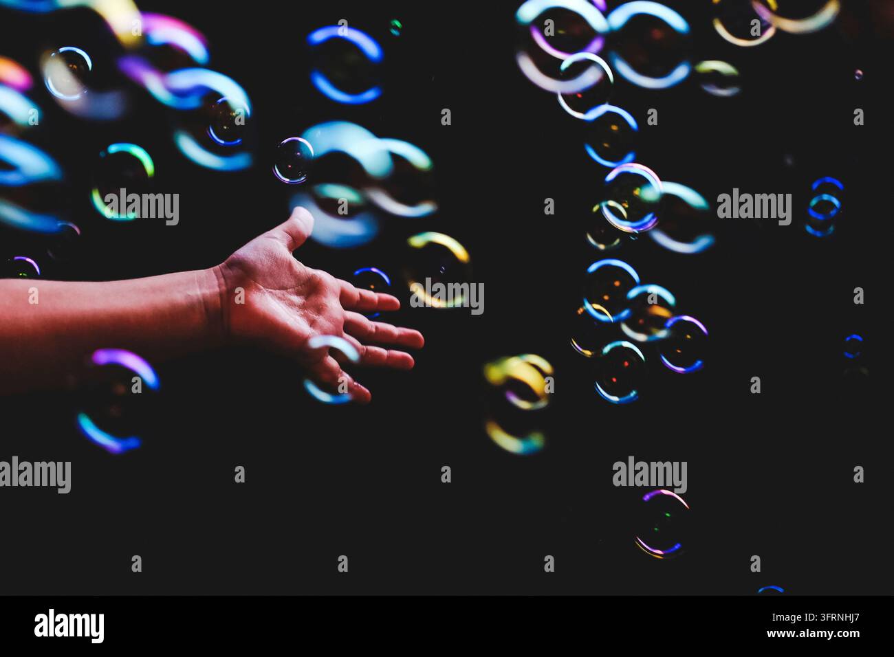 Bolle di sapone colorate volano su uno sfondo scuro e una mano umana cerca di catturarle Foto Stock