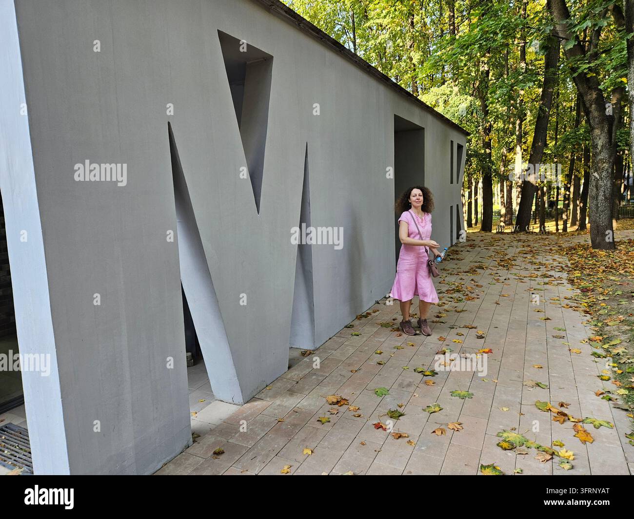 Donna in piedi davanti a un edificio con la lettera M su una toilette pubblica. Indossa un vestito rosa Foto Stock