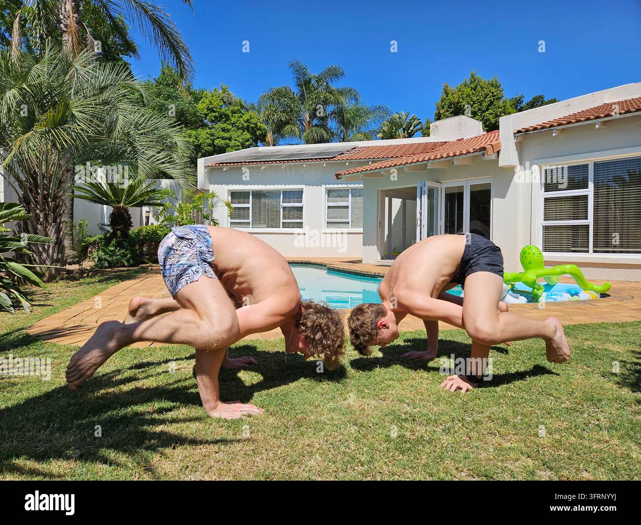 Due uomini stanno facendo yoga su un prato erboso. Uno di loro indossa pantaloncini con un disegno verde e bianco. padre e figlio in cortile con piscina a Johannesburg in Sudafrica (PR) Foto Stock