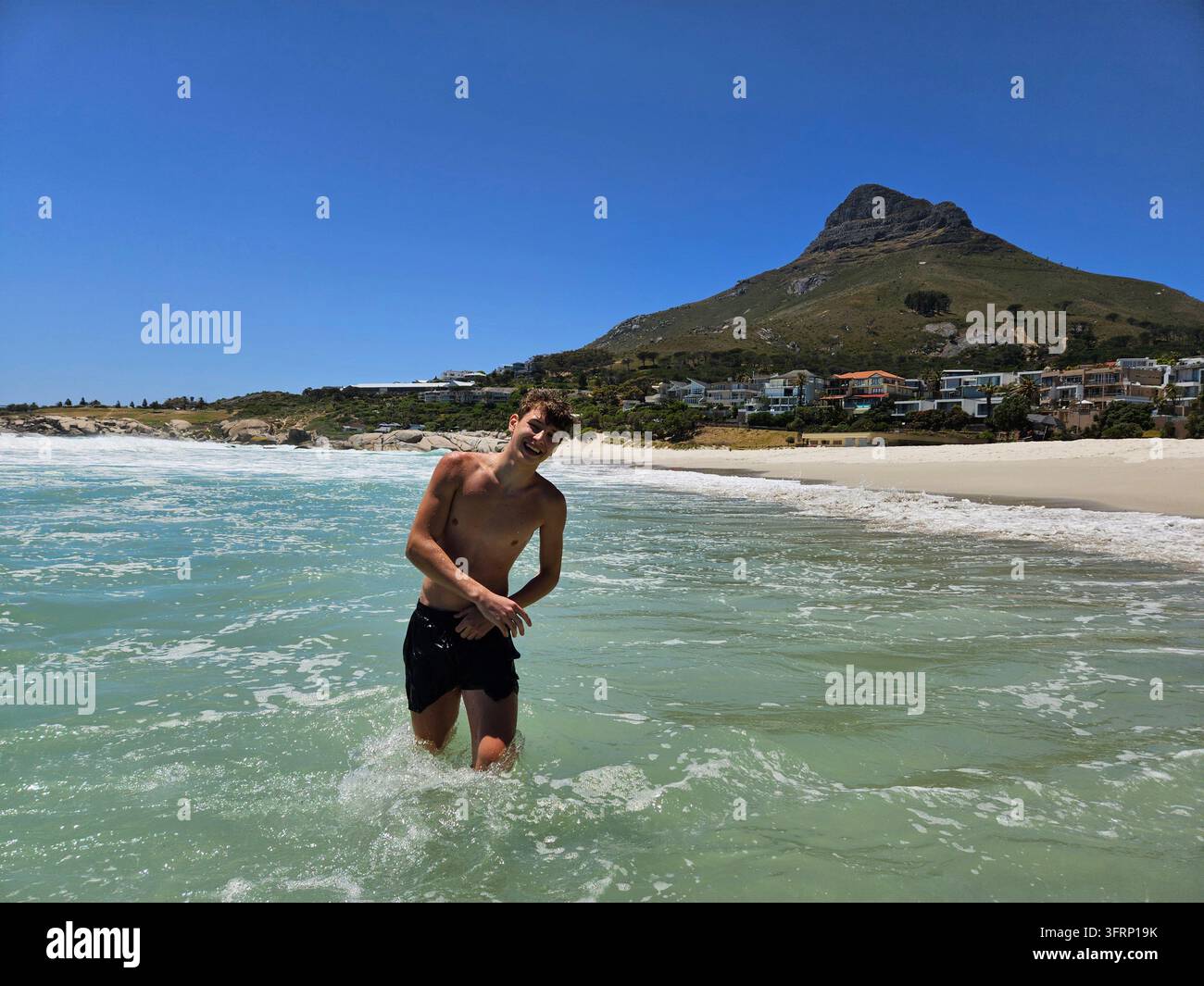 L'uomo è nell'oceano e indossa dei pantaloncini. Sta sorridendo. L'acqua è blu. Ci sono persone sulla spiaggia di Città del Capo in Sudafrica Foto Stock