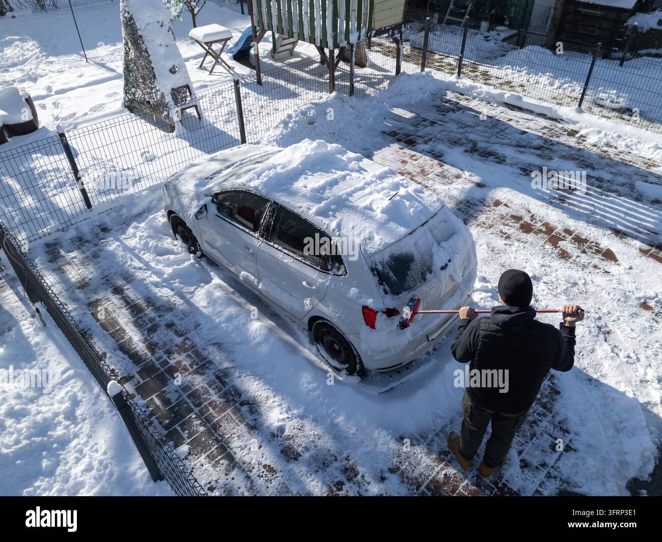 Una persona sta pulendo la neve da un'auto parcheggiata in uno spazio innevato durante la mattina d'inverno. Il terreno è coperto di neve. Foto Stock