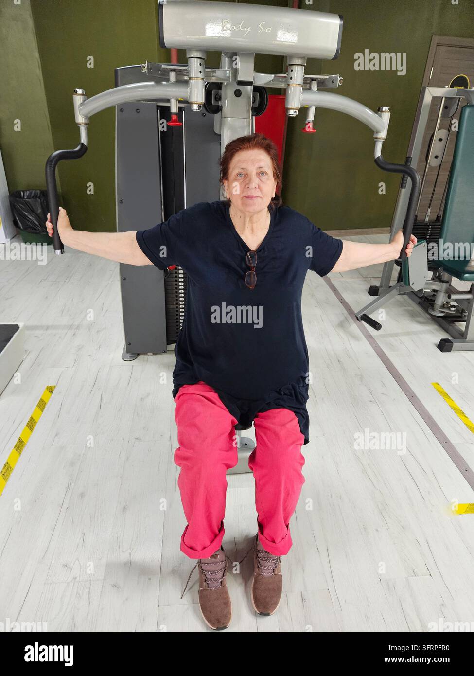 Una donna anziana è seduta su una macchina con un gatto sulla sua maglietta. Indossa pantaloni rosa e fa esercizi di allenamento sportivo Foto Stock