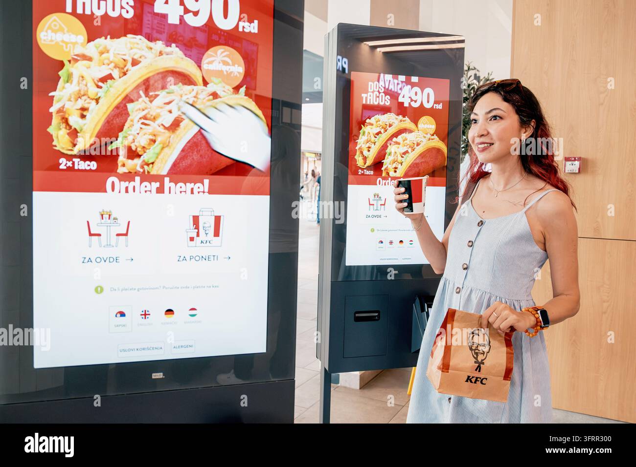 28 luglio 2026, Belgrado, Serbia: Giovane donna al chiosco di auto-ordinazione di KFC che tiene una borsa di marca e una bevanda, sorridente Foto Stock