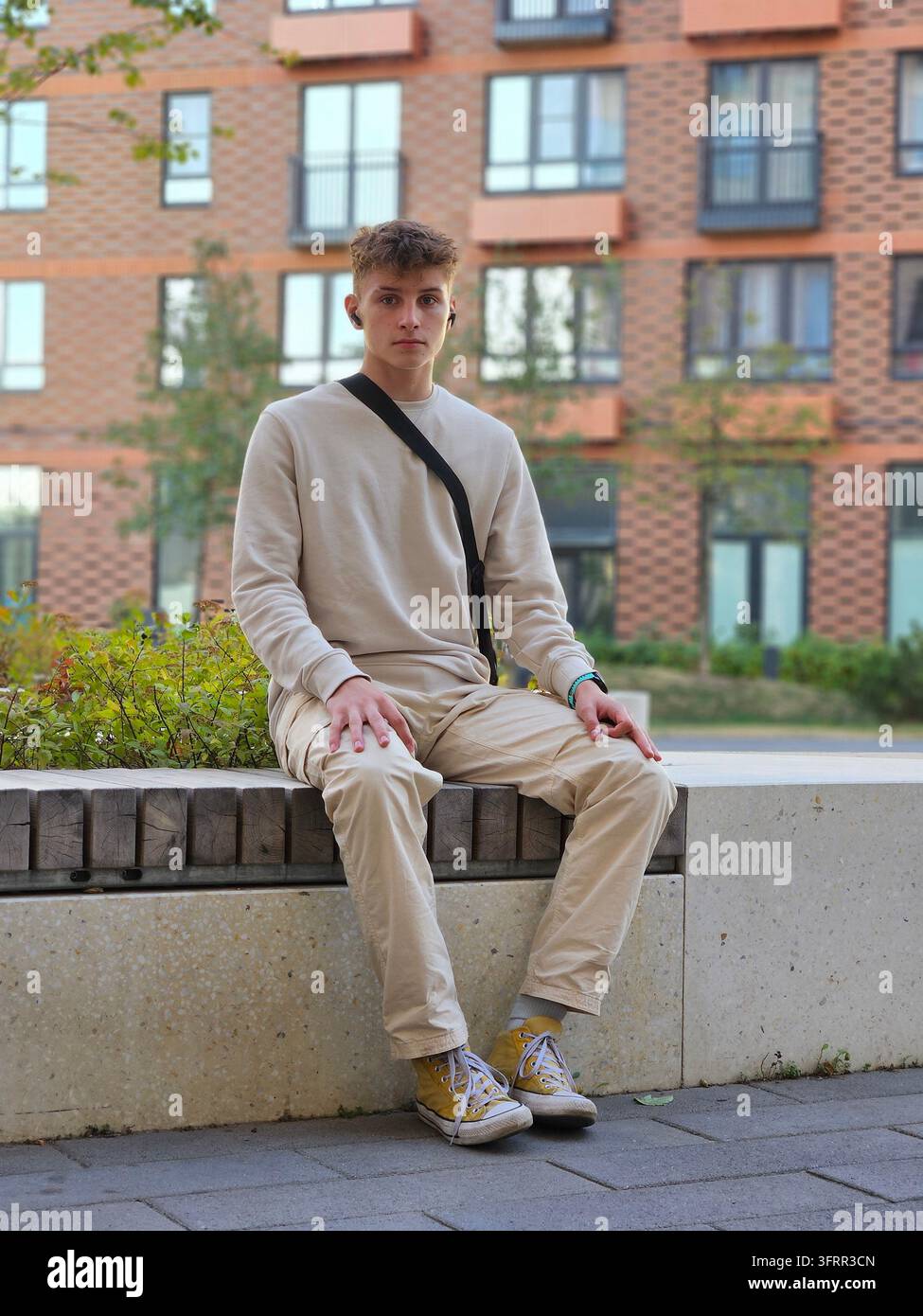 Un giovane uomo è seduto su una panchina indossando una camicia bianca e pantaloni marroni. Tiene in mano una borsa nera Foto Stock
