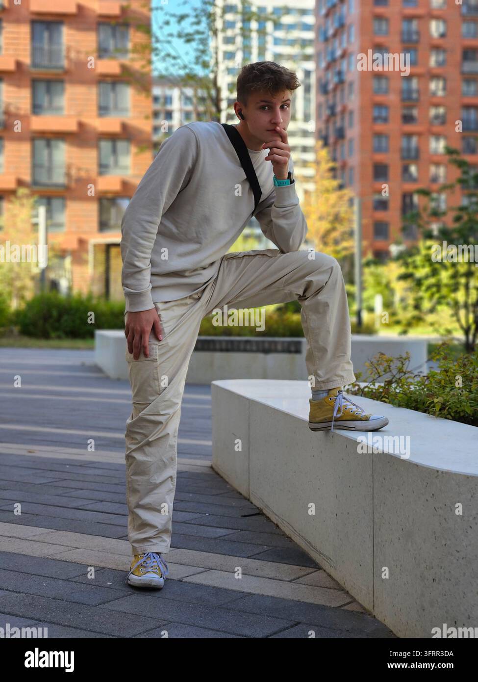 Uomo con camicia bianca e pantaloni marrone chiaro si appoggia a un muro. Indossa uno zaino nero ed è circondato da edifici alti Foto Stock