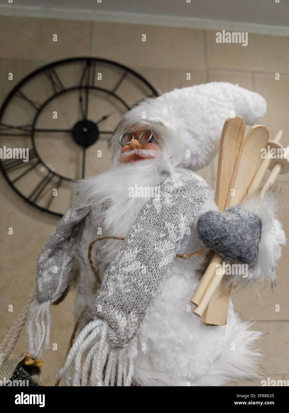 Babbo Natale bianco con una sciarpa grigia e occhiali che tiene due sci di legno. Un orologio è dietro di lui Foto Stock