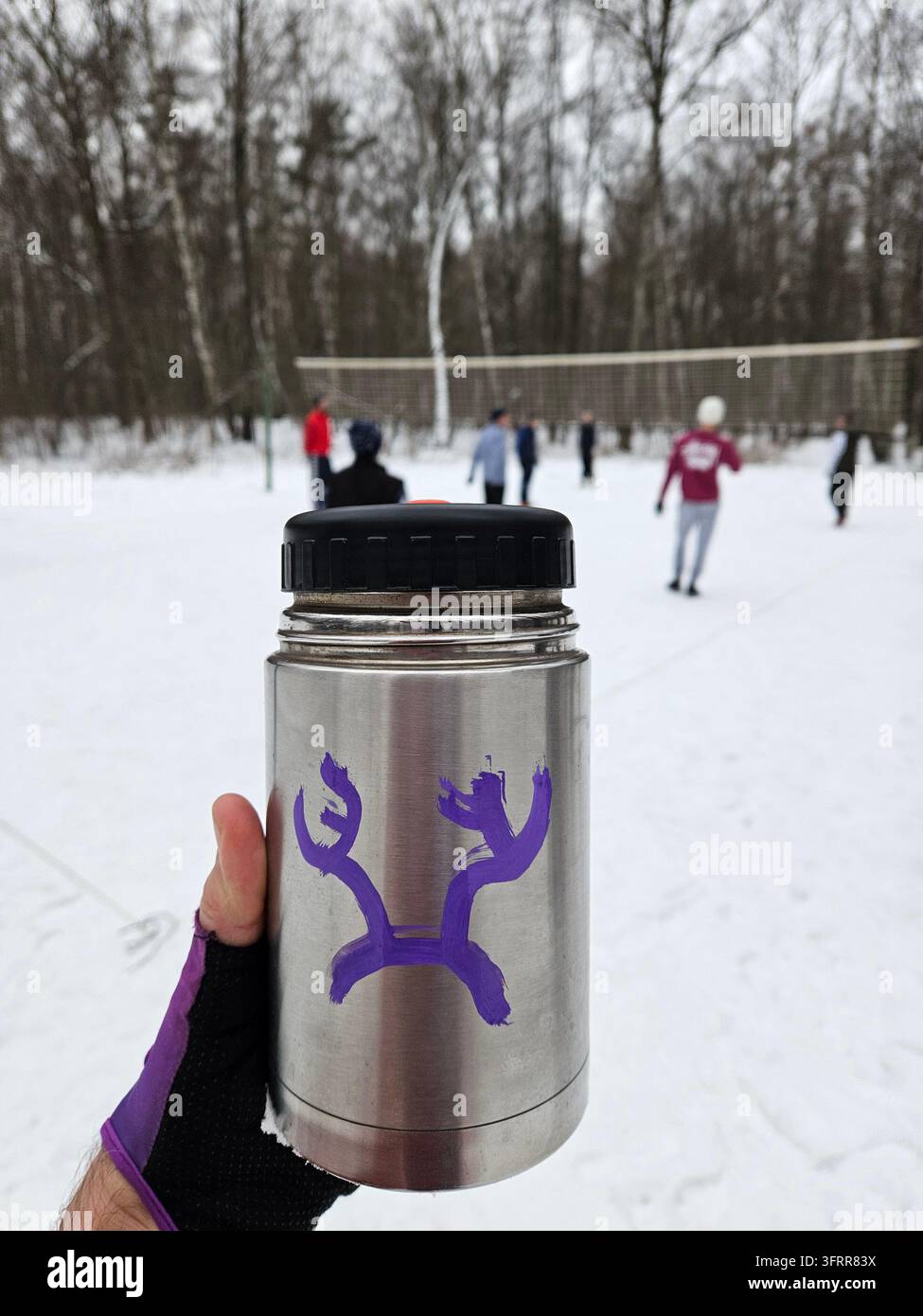 Una persona tiene un thermos argentato con un graffiti a forma di corno di cervo. Il thermos è viola e argento e ha un disegno di un cervo vicino a una partita di pallavolo in un parco invernale Foto Stock