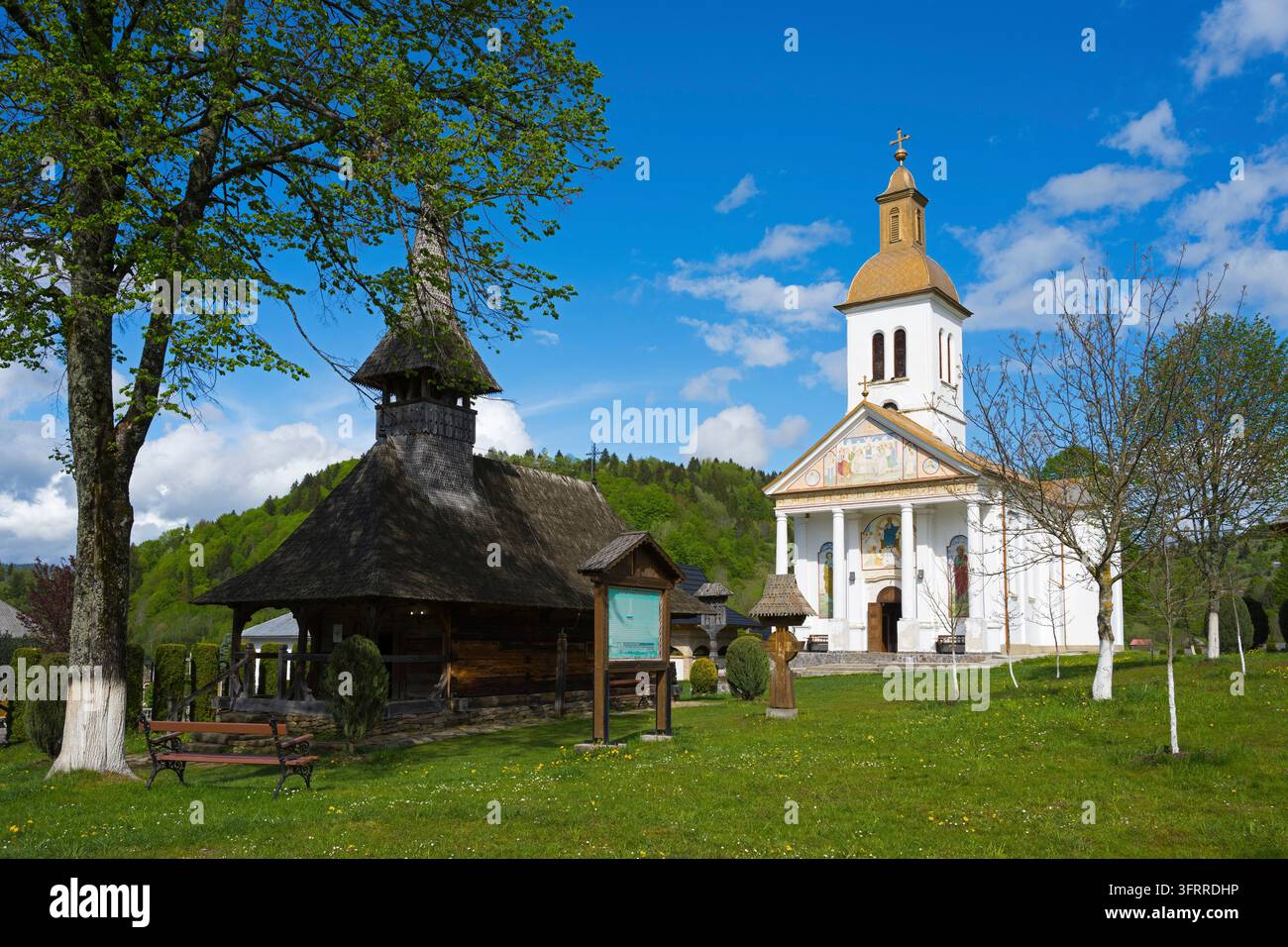 Una chiesa bianca e una chiesa storica in legno scura si ergono fianco a fianco in un prato soleggiato, chiesa in legno e chiesa, Manastirea Adormirea Maicii Domnulu Foto Stock