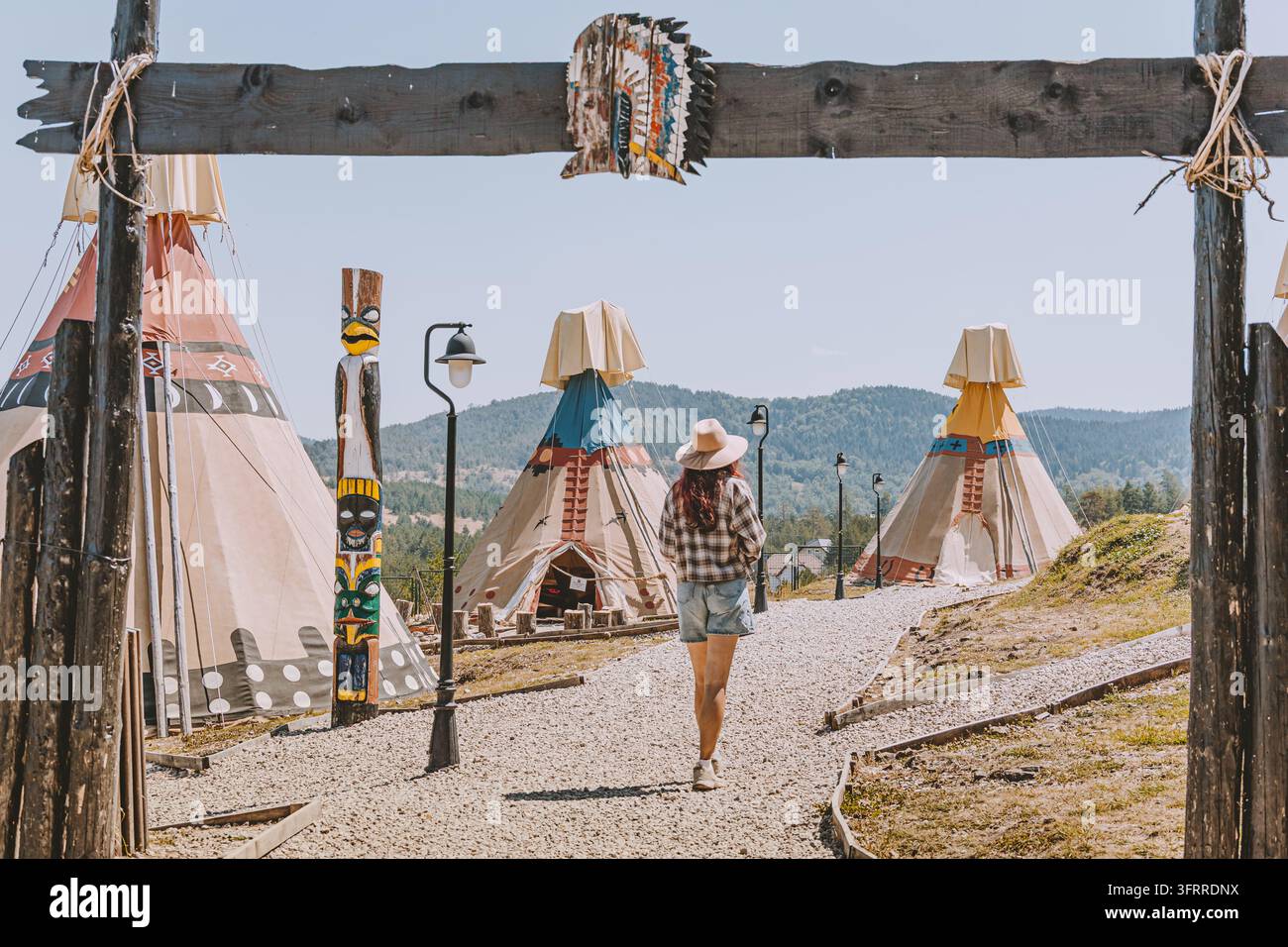 17 agosto 2026, Zlatibor, parco a tema El Paso, Serbia: donna che attraversa un arco in legno verso un campo di teepee con palo totem e sfondo montano. Foto Stock
