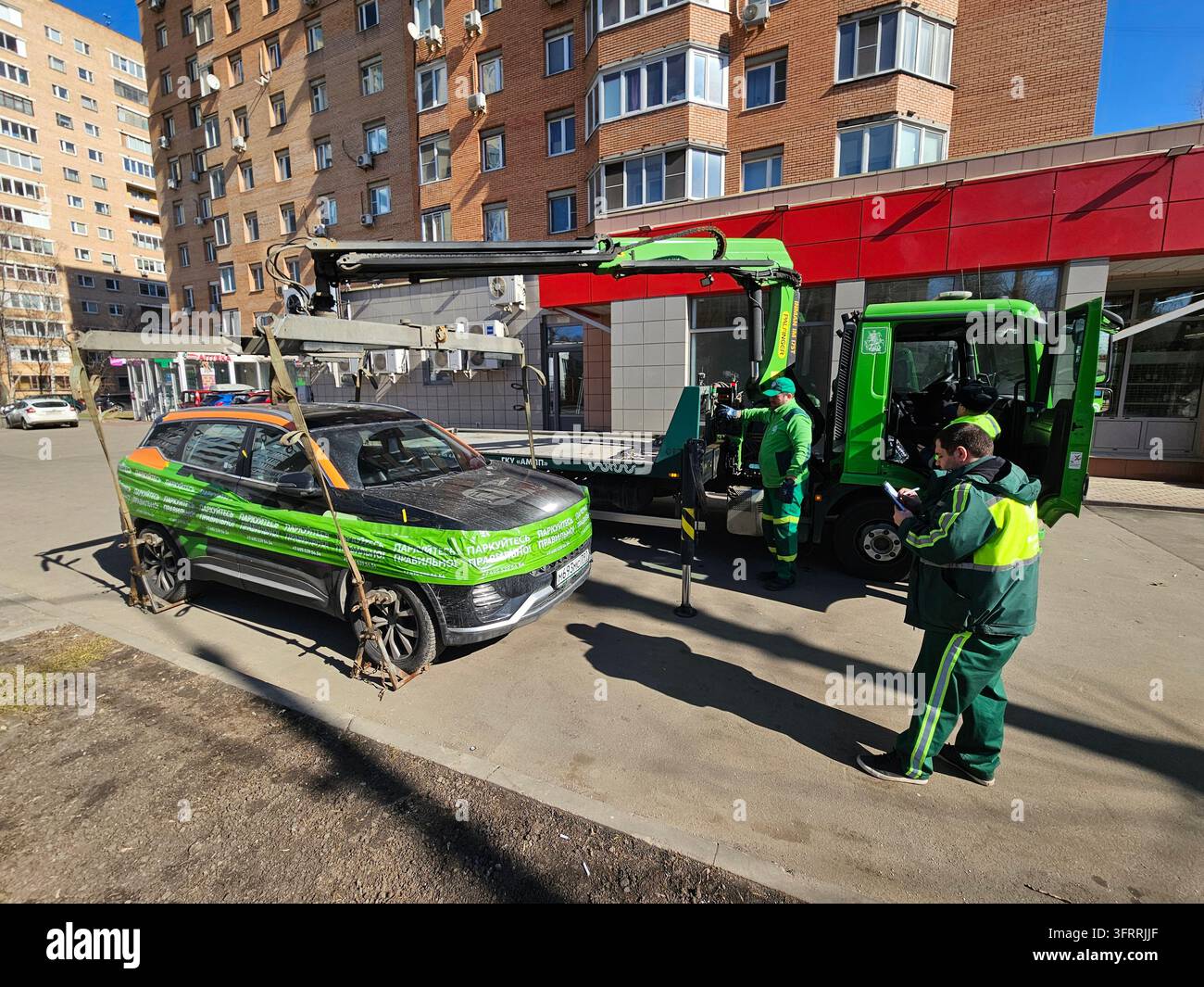 Mosca - 19 marzo 2025: Un'auto con nastro verde sul marciapiede viene sollevata da un camion gru e da un trasgressore del parcheggio. Un uomo in un giubbotto verde è in piedi accanto all'auto Foto Stock