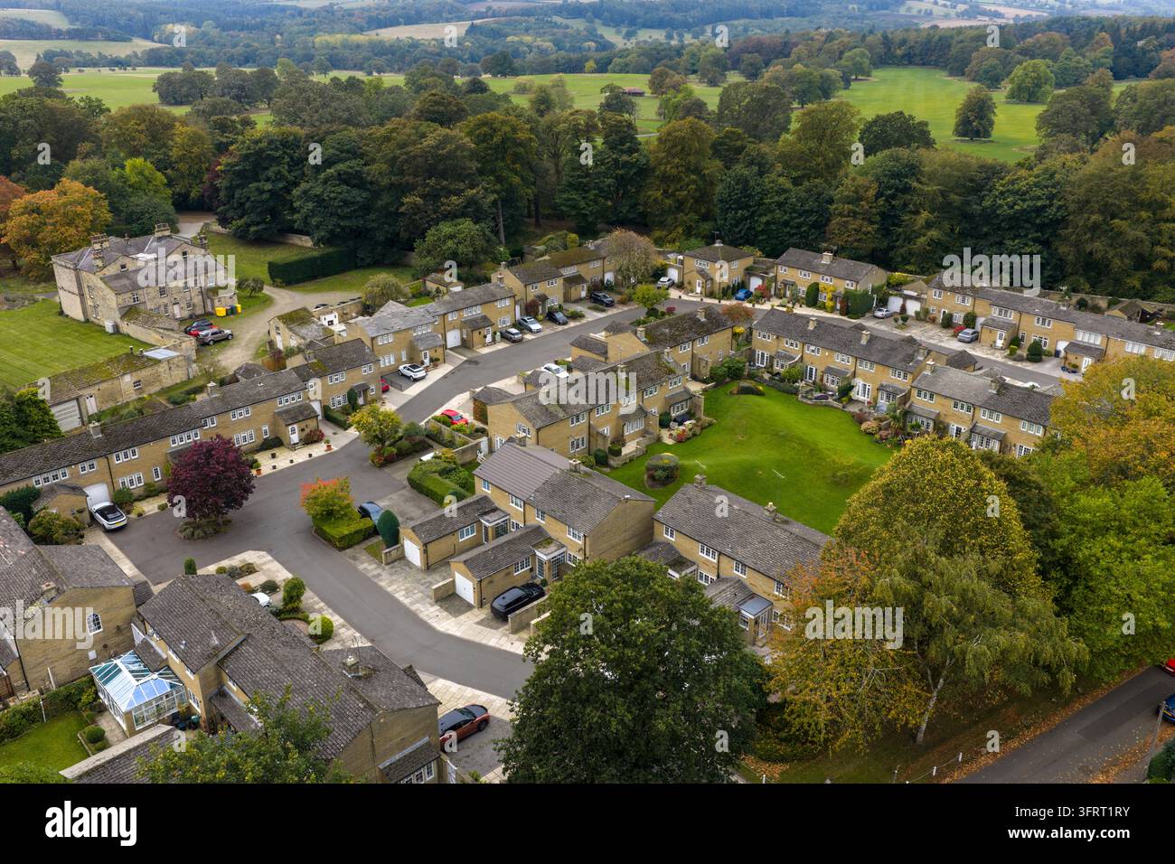 Fotografia a drone che si affaccia su un tranquillo quartiere residenziale a Harewood con file di case in pietra, vialetti, veicoli parcheggiati, prati e bordo boscoso Foto Stock