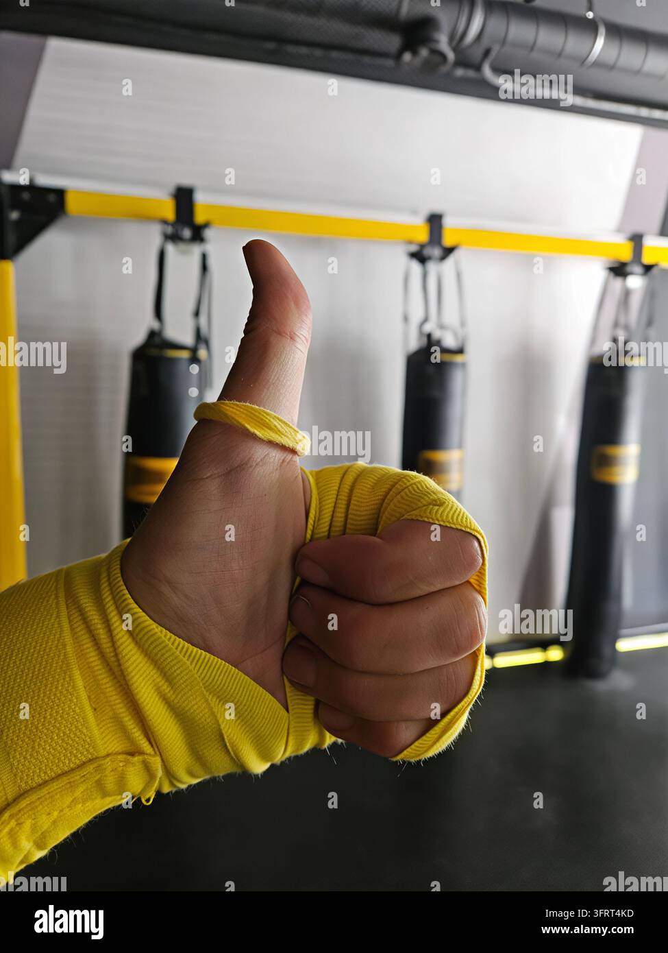 Una persona fa un gesto di pollice alzato con un nastro giallo sulla mano. Il pollice alzato è situato al centro della mano, una fila di sacchi da boxe è appesa al soffitto. I sacchi sono neri e gialli Foto Stock