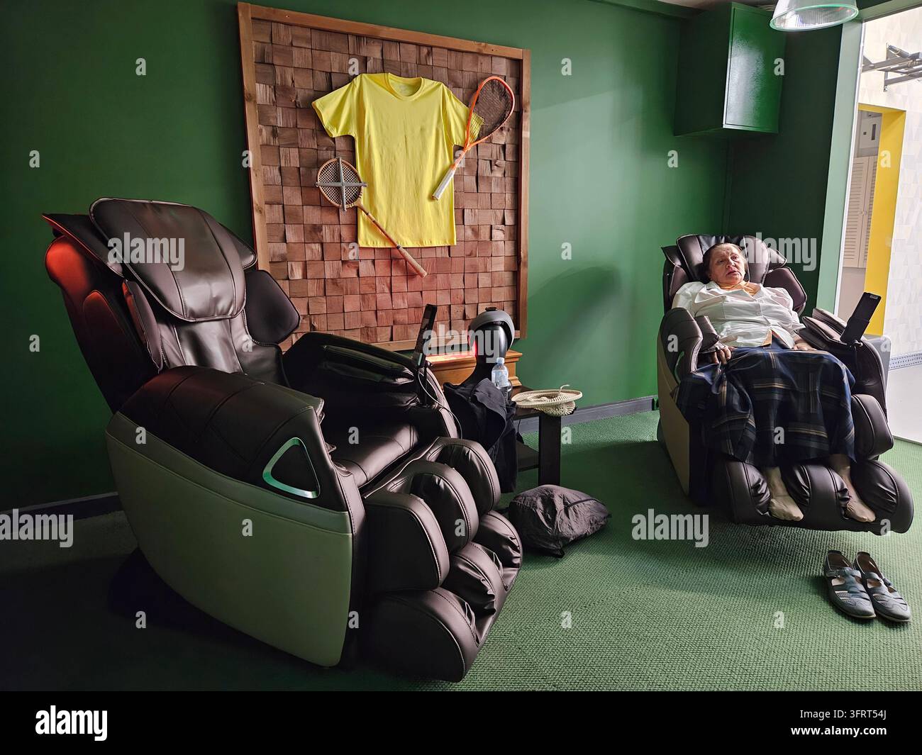Una donna anziana è seduta su una sedia da massaggio con i piedi sollevati. C'è un muro con una camicia sopra nella sala relax del complesso sportivo Foto Stock