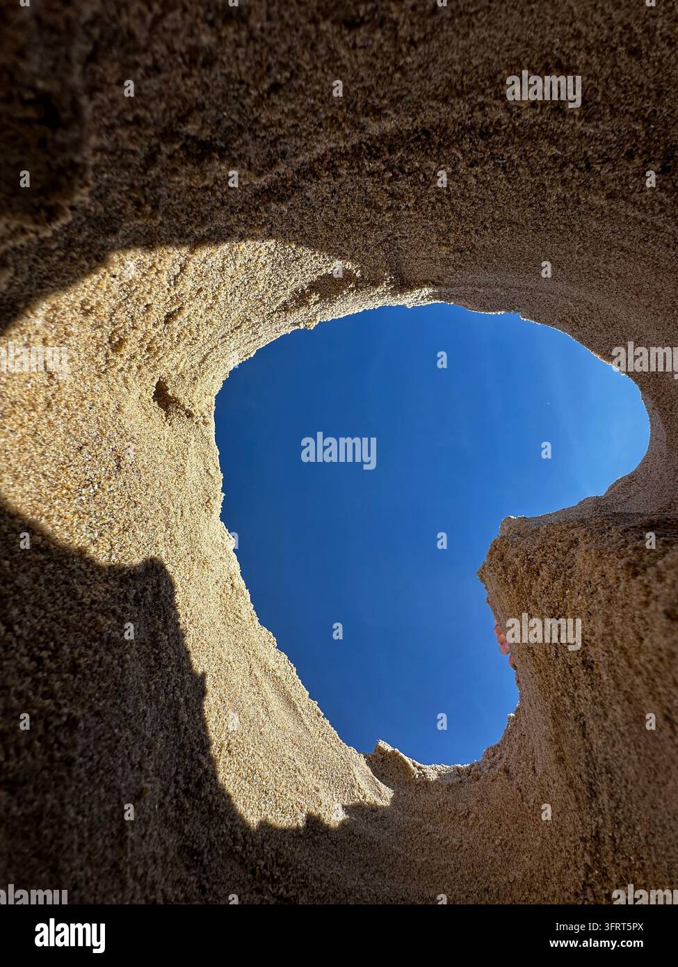 Vista ad angolazione bassa che guarda verso il cielo azzurro e le nuvole dall'interno di un buco a forma di cuore scavato nella sabbia su una spiaggia o sul terreno - Immagine stock catturata con smartphone