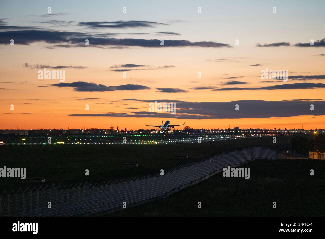 Aereo passeggeri che si eleva nel cielo colorato della sera poco dopo il decollo, sopra la pista illuminata e la recinzione perimetrale dell'aeroporto. Foto Stock
