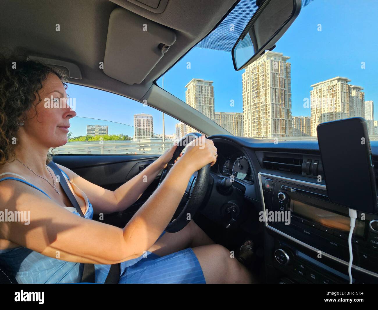 Una donna guida un'auto con una città sullo sfondo. Sta sorridendo e si sta godendo il suo viaggio Foto Stock