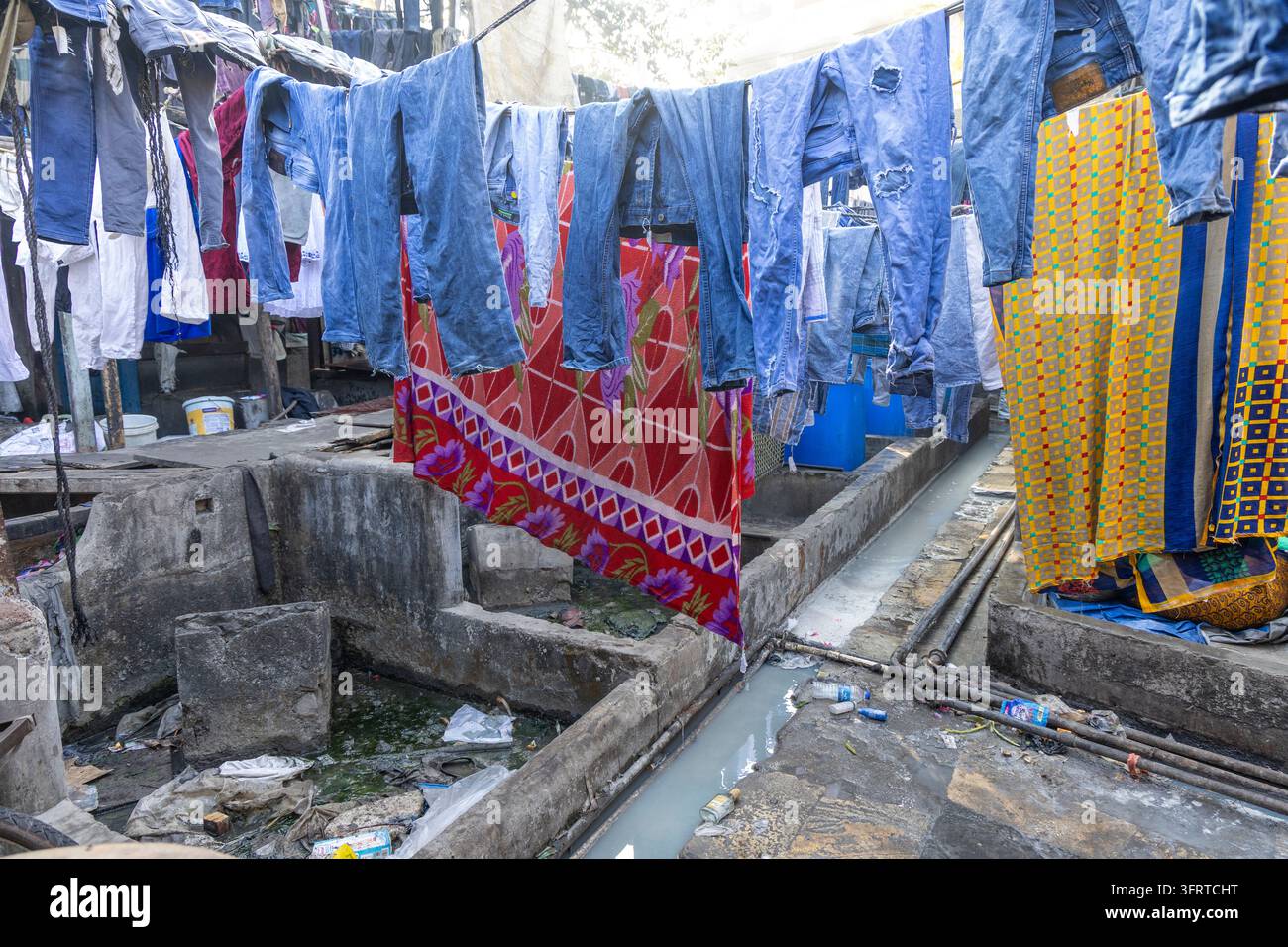 Jeans di denim e tessuti colorati appesi ad asciugare sopra vasche di lavaggio in cemento vuote e un canale a Dhobi Ghat, Maharashtra, Mumbai, India Foto Stock