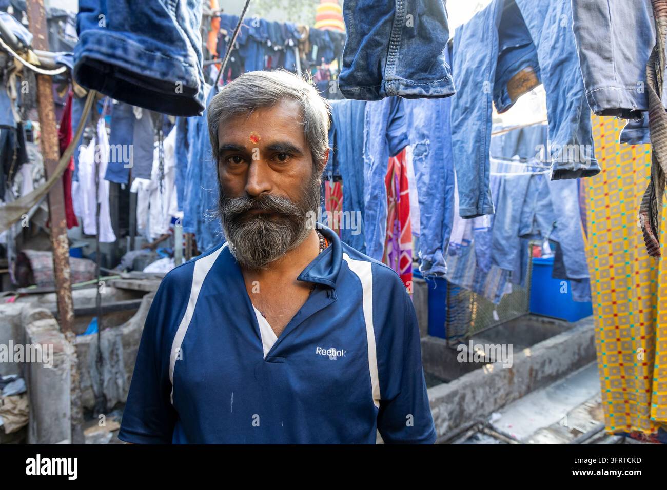 Ritratto di un uomo barbuto con un tilak rosso sulla fronte in piedi tra jeans blu appesi ad asciugare a Dhobi Ghat, Maharashtra, Mumbai, India Foto Stock