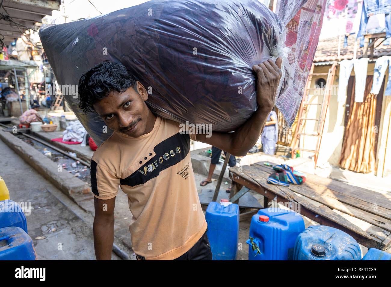 Uomo sorridente che porta un enorme sacco di biancheria sulla spalla attraverso un vicolo stretto con taniche blu a Dhobi Ghat, Maharashtra, Mumbai, India Foto Stock
