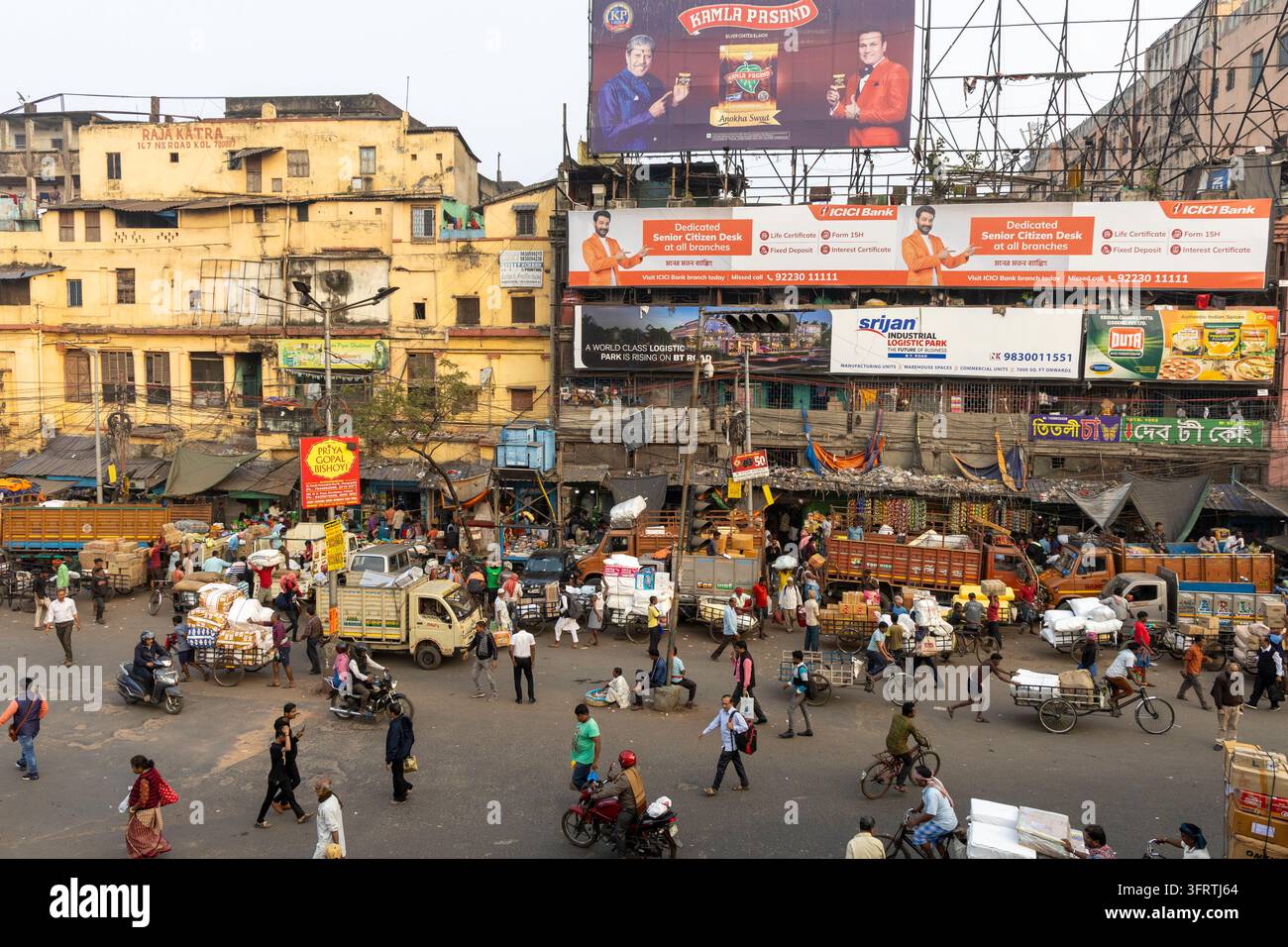 Vista ad angolazione alta di un ampio incrocio pieno di camion, carri a mano, risciò e persone che camminano davanti ad edifici gialli logorati e cartelloni pubblicitari, Noi Foto Stock