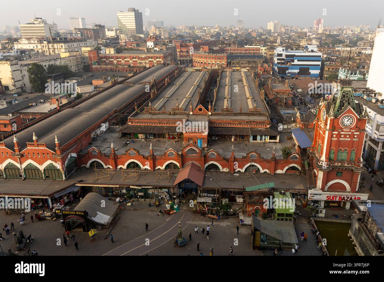 Vista ad angolazione alta di un complesso mercatale storico in mattoni rossi con facciate ad arco, una torre dell'orologio e lunghe hall coperte circondate da edifici cittadini, Bengala Occidentale Foto Stock