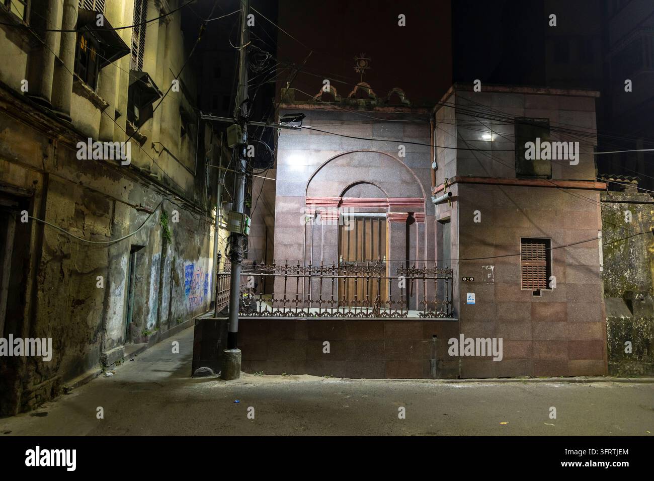Vicolo lastricato vuoto con edifici coloniali erositi, cavi aerei e una piccola struttura con pannelli ad arco che brillano sotto i riflettori, Bengala Occidentale, K Foto Stock