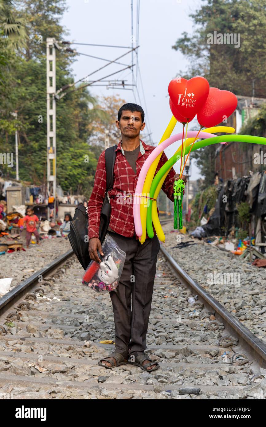 Venditore ambulante con una camicia a scacchi rossi e una borsa a tracolla si trova tra i binari portando palloncini ritorti e un sacchetto di plastica, Bengala Occidentale, Foto Stock