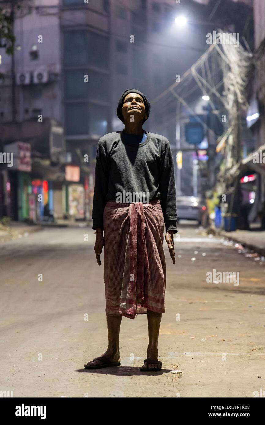 Lavoratore solitario in un maglione scuro si trova con gli occhi rivolti verso i lampioni sopra una strada deserta fiancheggiata da negozi chiusi e alti edifici, Bengala Occidentale Foto Stock