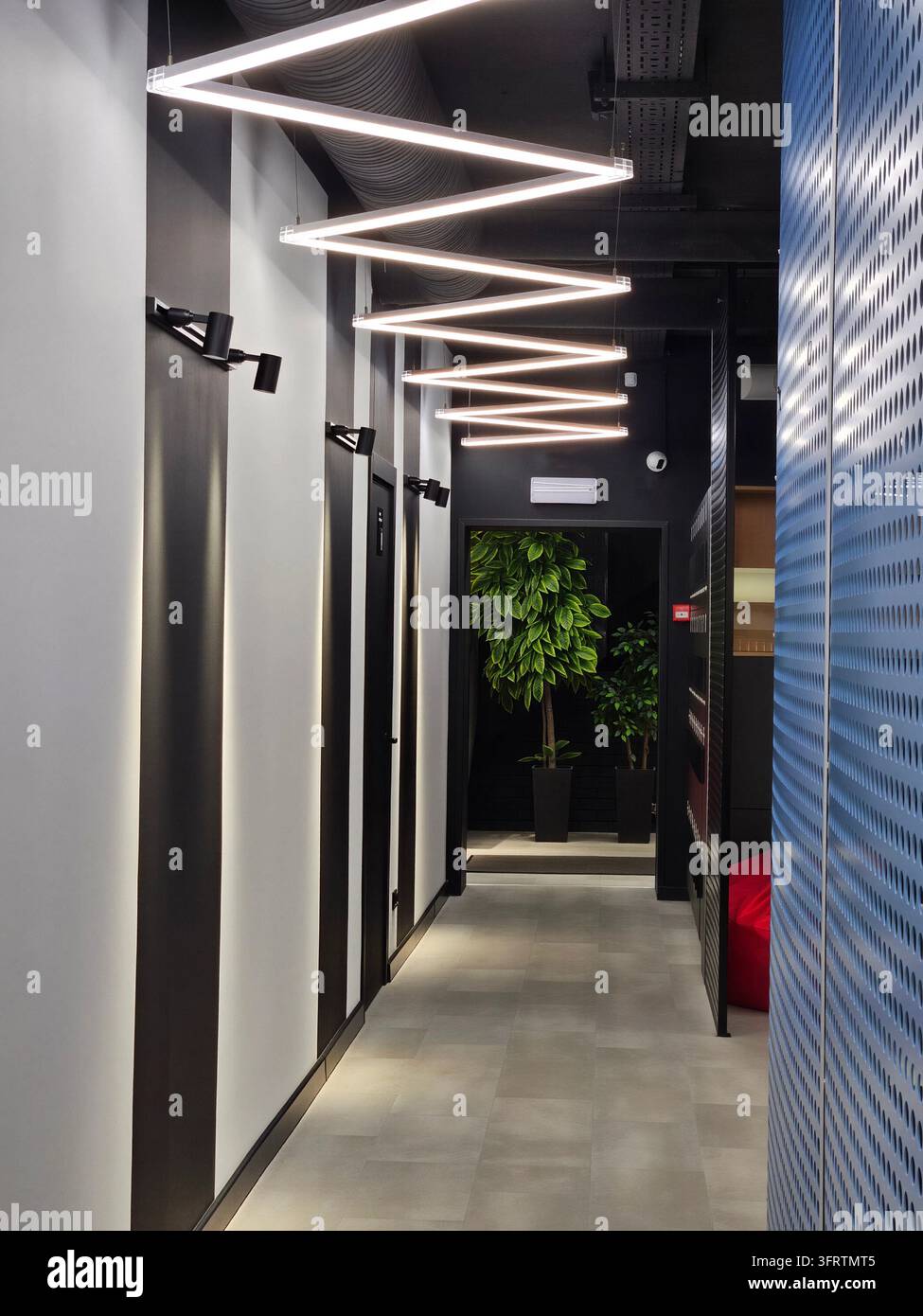 Corridoio moderno con illuminazione a LED a zigzag. Pannelli scuri contrastano con pareti bianche. Una pianta verde aggiunge un tocco naturale allo spazio. Il pavimento è in cemento lucido. Un pannello perforato blu delimita il lato destro Foto Stock