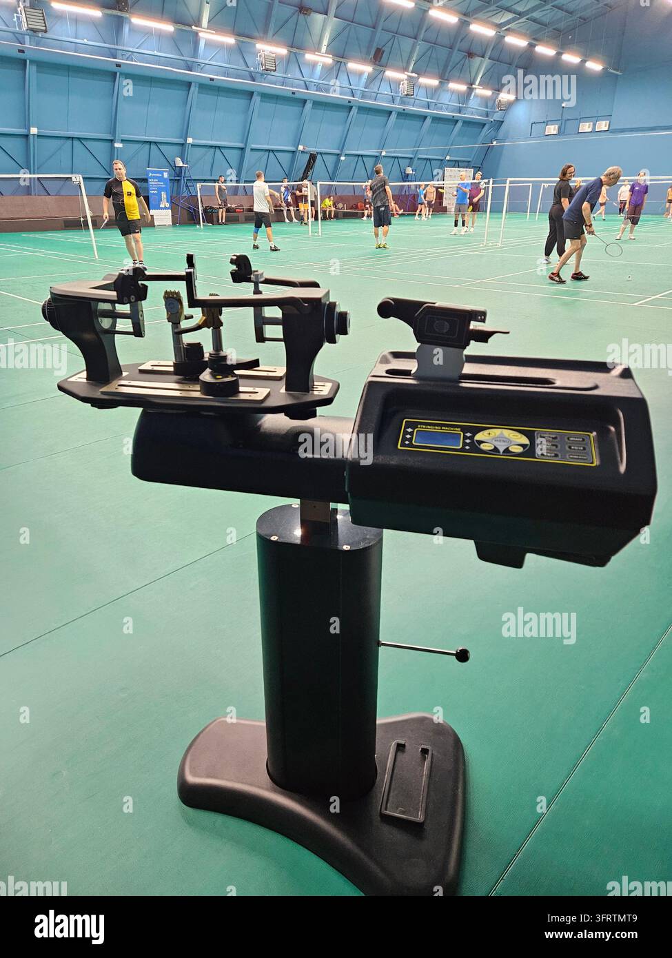 Macchina professionale per stringere le racchette da tennis su campo al coperto. I giocatori si allenano sullo sfondo. Pavimentazione verde, soffitto alto, illuminazione brillante. La macchina ha un display digitale e bracci regolabili Foto Stock