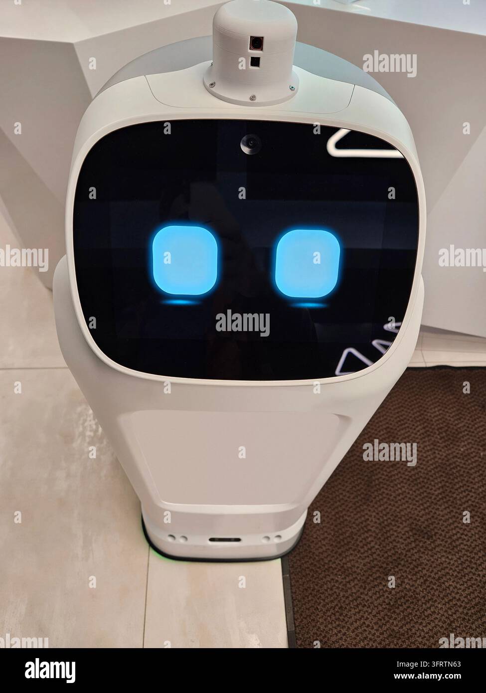 Robotto moderno con occhi blu luminosi in piedi al chiuso. L'involucro bianco presenta un design elegante e arrotondato. Il pannello frontale mostra uno schermo digitale con due cerchi illuminati Foto Stock