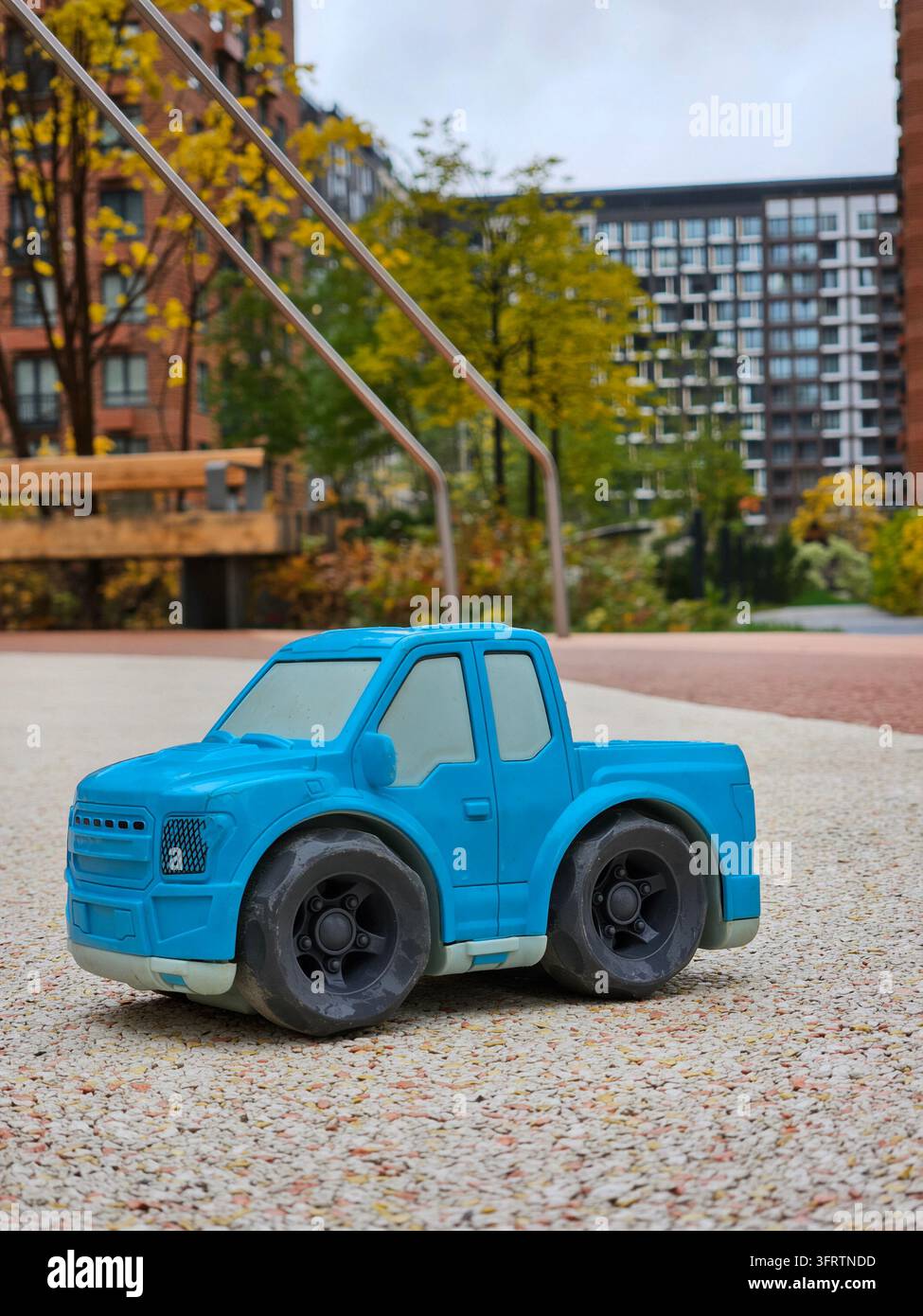 Camioncino giocattolo di plastica blu si trova su un sentiero di ghiaia. Lo sfondo presenta edifici urbani e alberi autunnali. La messa a fuoco morbida aggiunge profondità alla scena. Il colore blu brillante attira immediatamente l'attenzione Foto Stock