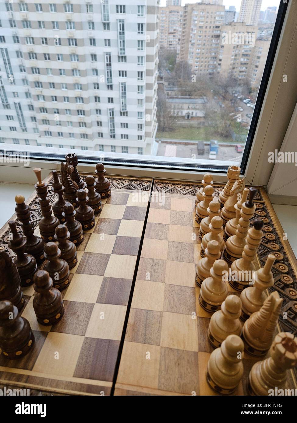 Scacchiera pronta per la partita, vista dall'alto. Pedine di legno disposte nelle posizioni di partenza. Paesaggio urbano visibile attraverso una grande finestra dietro. La luce naturale illumina la scacchiera e le pedine Foto Stock