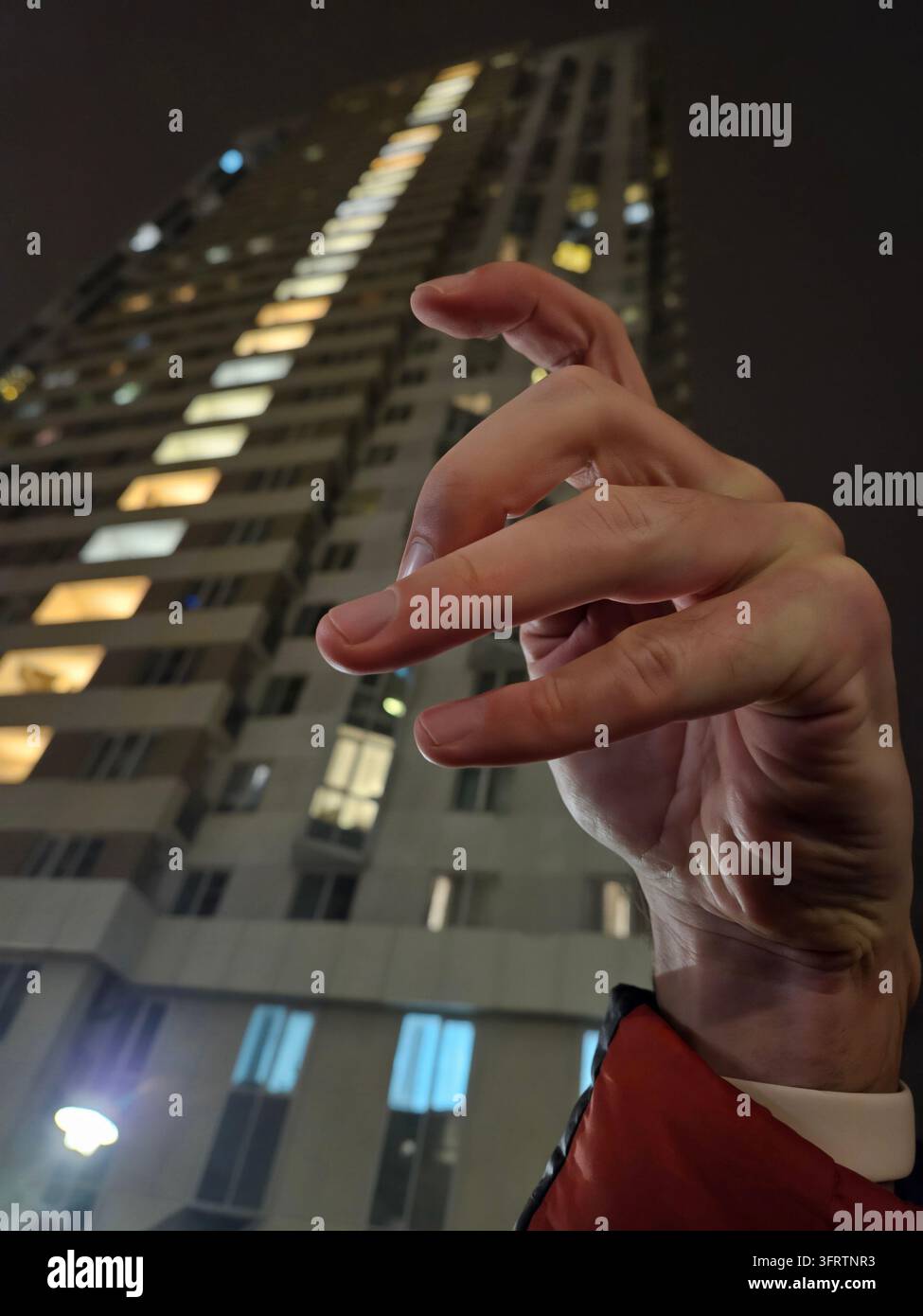 Paesaggio urbano notturno con grattacielo illuminato. Gesti delle mani in primo piano, dita leggermente curvate. La persona indossa una manica rossa, in piedi vicino all'edificio. Le luci urbane si riflettono nei vetri delle finestre Foto Stock