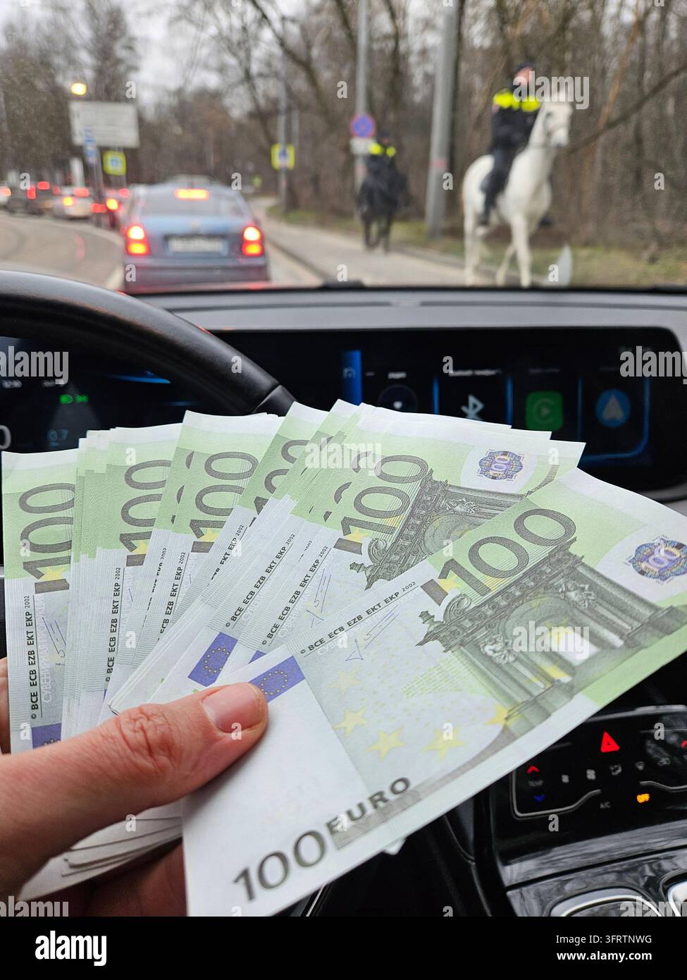 Una mano tiene banconote in euro all'interno di un'auto. Un agente di polizia cavalca un cavallo accanto alla strada. Il conducente osserva il traffico davanti a lui dal cockpit. Valuta sventolata vicino al volante. Il cruscotto del veicolo mostra controlli digitali Foto Stock