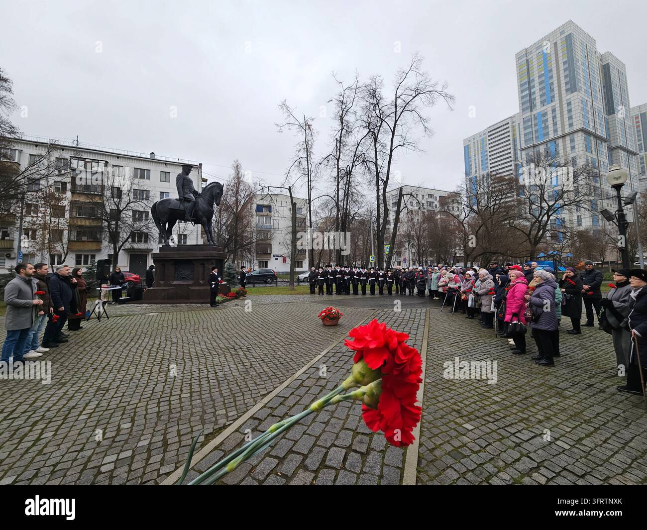 Mosca - 05 dic. 2025: Garofani rossi accovacciati giacciono sul terreno di ciottoli. La statua della figura montata Rokossovsky si erge al centro della piazza. Una folla si raduna in silenzio rispettoso attorno al monumento Foto Stock