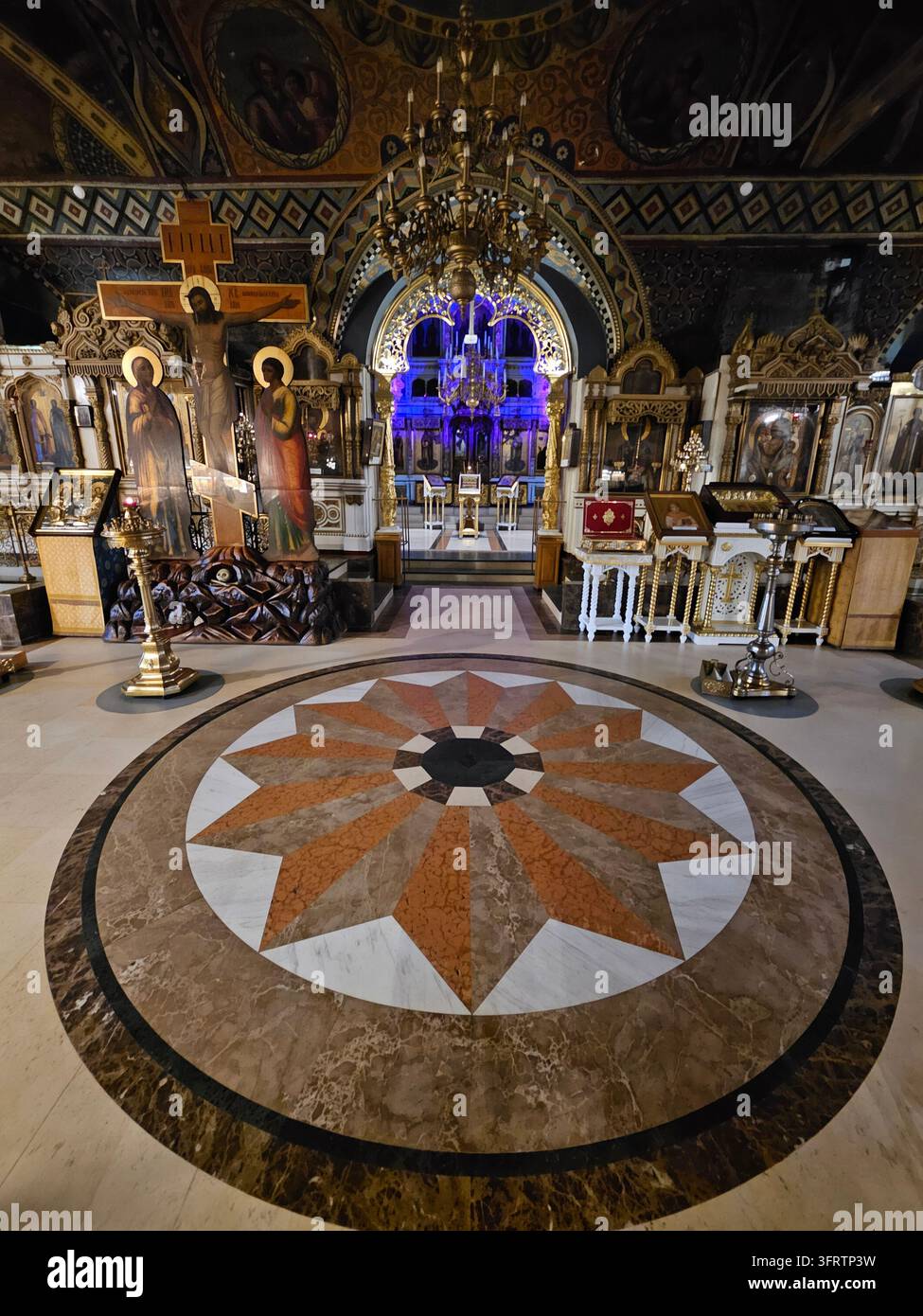 Mosca – 18 dic. 2025: Interno di chiesa serba riccamente decorato con pavimento a mosaico ornato. Lampadari dorati pendono sopra altari sacri. Icone religiose vibranti adornano pareti e piedistalli Foto Stock