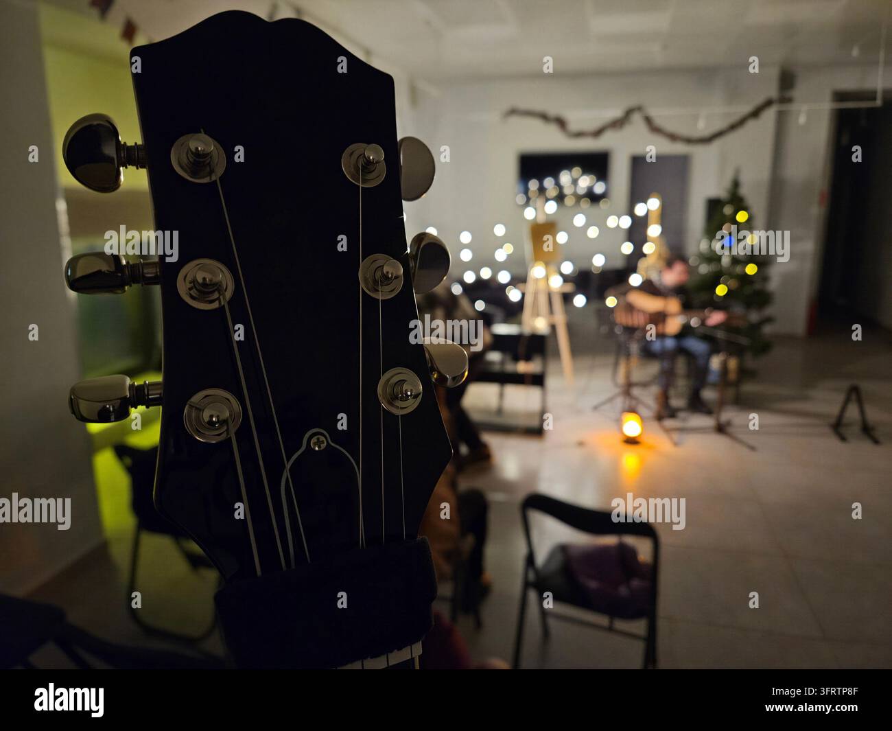 Il capo della chitarra domina lo sfondo, lo sfondo è sfocato dolcemente. Una stanza accogliente risplende con luci calde delle feste. Una persona suona la chitarra in un ambiente festivo lontano. Un albero si trova accanto al musicista seduto Foto Stock