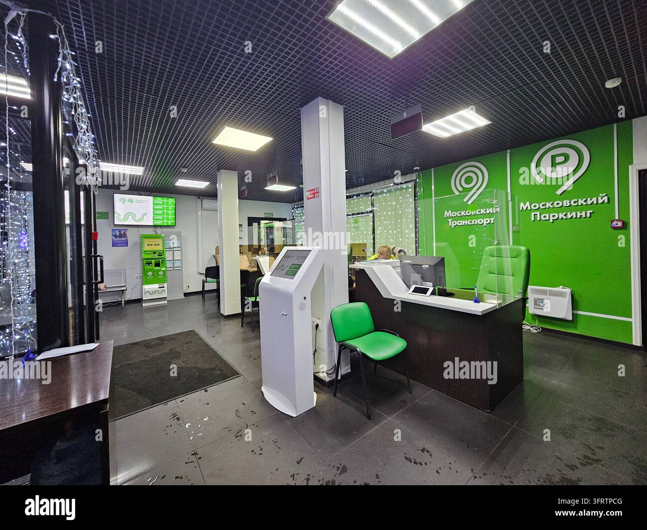 Mosca - 26 dic. 2025: Parcheggio ufficio moderno con penalità per spazio di guida scarso, marchio verde e banco di ricezione. Chiosco bianco e sedia verde completano il layout pulito. Piastrelle per soffitto e illuminazione a incasso Foto Stock