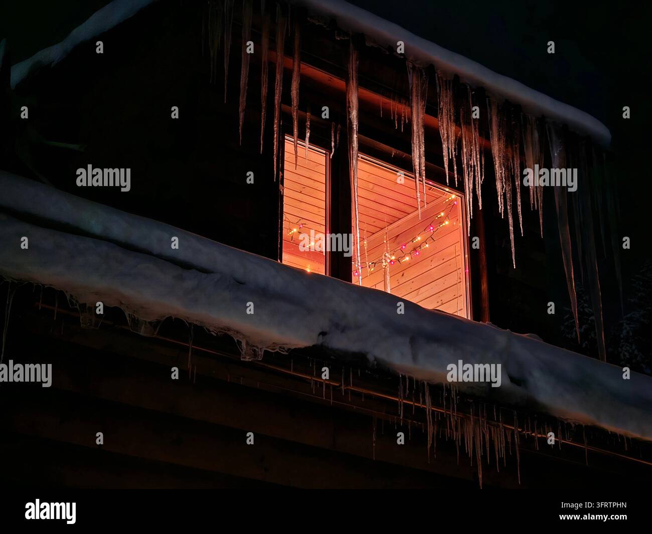 Tetto coperto di neve brilla calorosamente dall'interno. Lunghe ghiaccioli pendono dalle grondaie di notte. La cornice di legno della finestra emana una luce soffusa e dorata. L'esterno oscurato contrasta con l'interno accogliente (PR) Foto Stock
