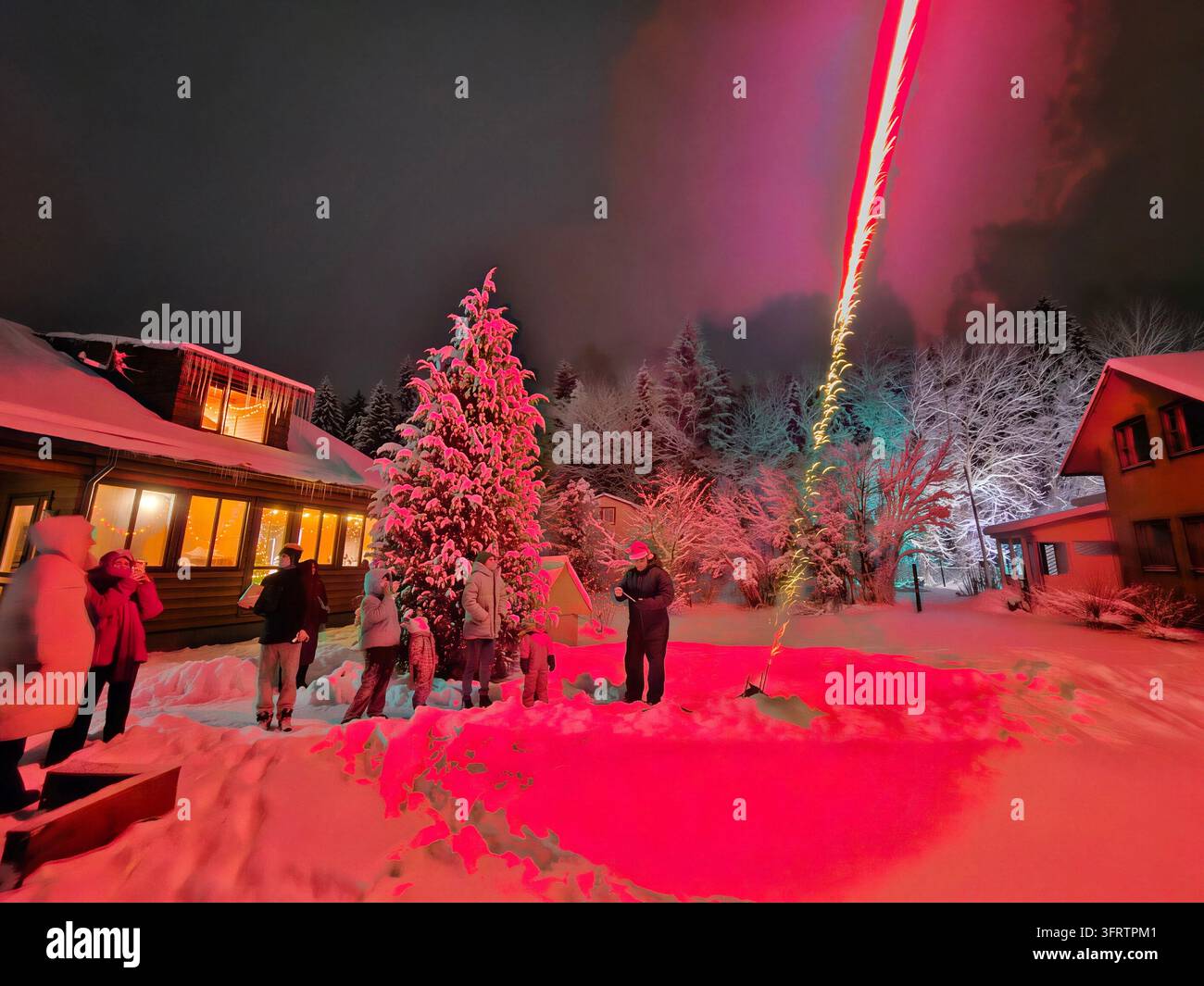 Notte nevosa con luci festive e raggi laser. Un gruppo si raduna vicino a un pino illuminato. Gli edifici brillano di un caldo bagliore dietro il terreno coperto di neve. Un raggio laser rosso taglia il cielo scuro Foto Stock