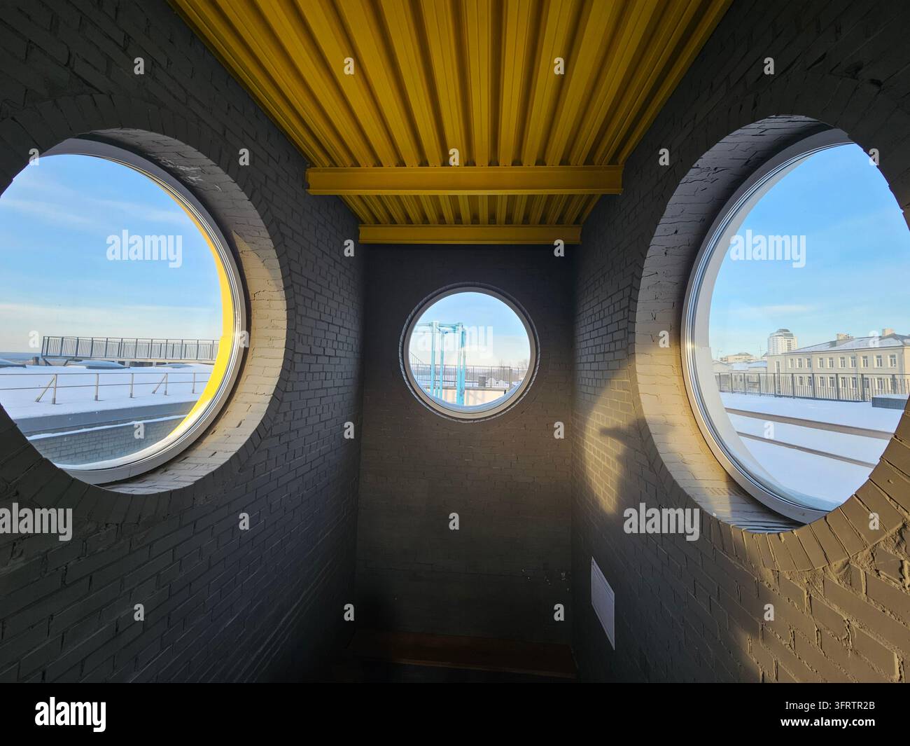 SAMARA - 14 GEN 2026: Finestre circolari incorniciano viste sul lungomare innevato. Il soffitto giallo contrasta con le pareti di mattoni grigi. La luce del sole mette in risalto le superfici ruvide dall'interno. Paesaggio urbano visibile attraverso rotonde Foto Stock