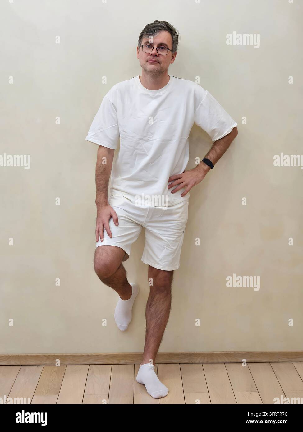 Uomo in piedi davanti a un muro semplice con indumenti bianchi. Indossa occhiali e calzini bianchi. Una mano poggia sul fianco, l'altra sul ginocchio. Lo sfondo semplice enfatizza la posa casual e rilassata Foto Stock