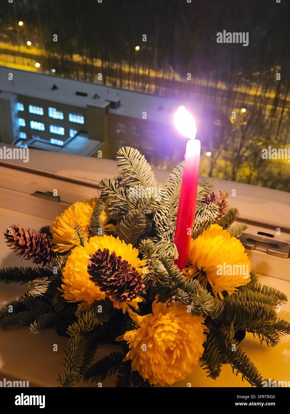 Calda luce di candela accanto a fiori gialli e rami di pino. Disposizione festiva appoggiata sul davanzale di notte. Alberi e edifici innevati visibili sullo sfondo Foto Stock