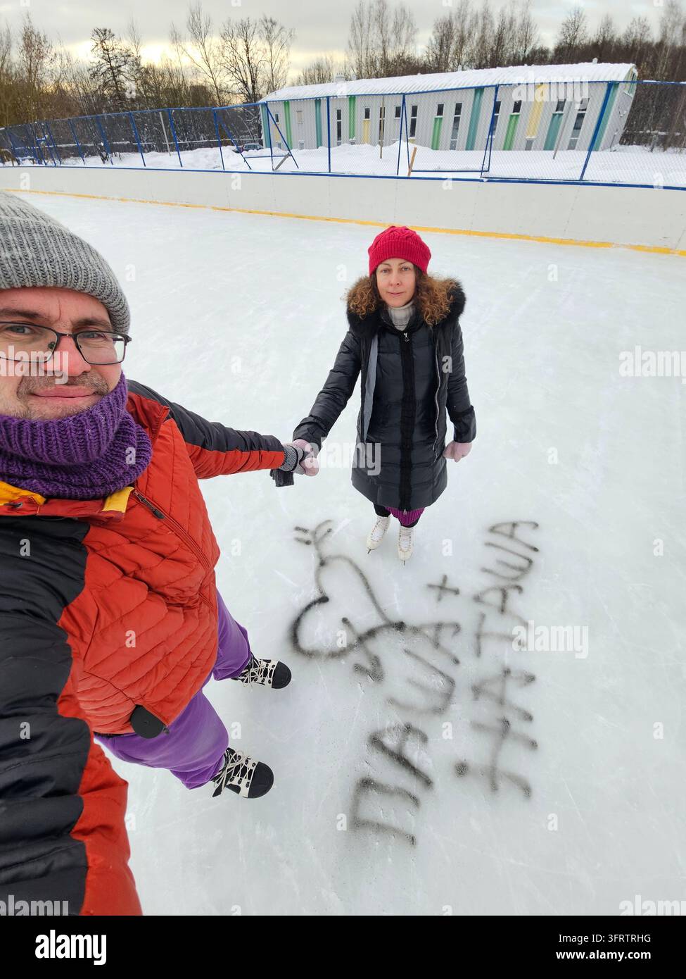 Due persone si tengono per mano su una pista di ghiaccio. Un cuore e dei nomi sono disegnati nella neve. Indossano cappotti invernali e cappelli. Una persona scatta un selfie con una fotocamera. La neve copre il terreno e l'area circostante Foto Stock