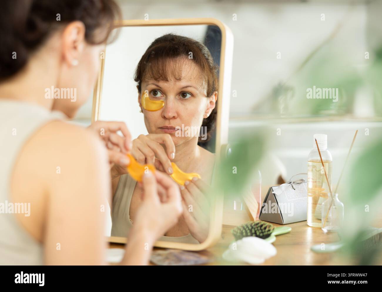 Donna sulla quarantina seduta davanti a uno specchio da trucco in una stanza del mattino ben illuminata, utilizza cerotti occhi al collagene dorati per idratare e ridurre le rughe Foto Stock