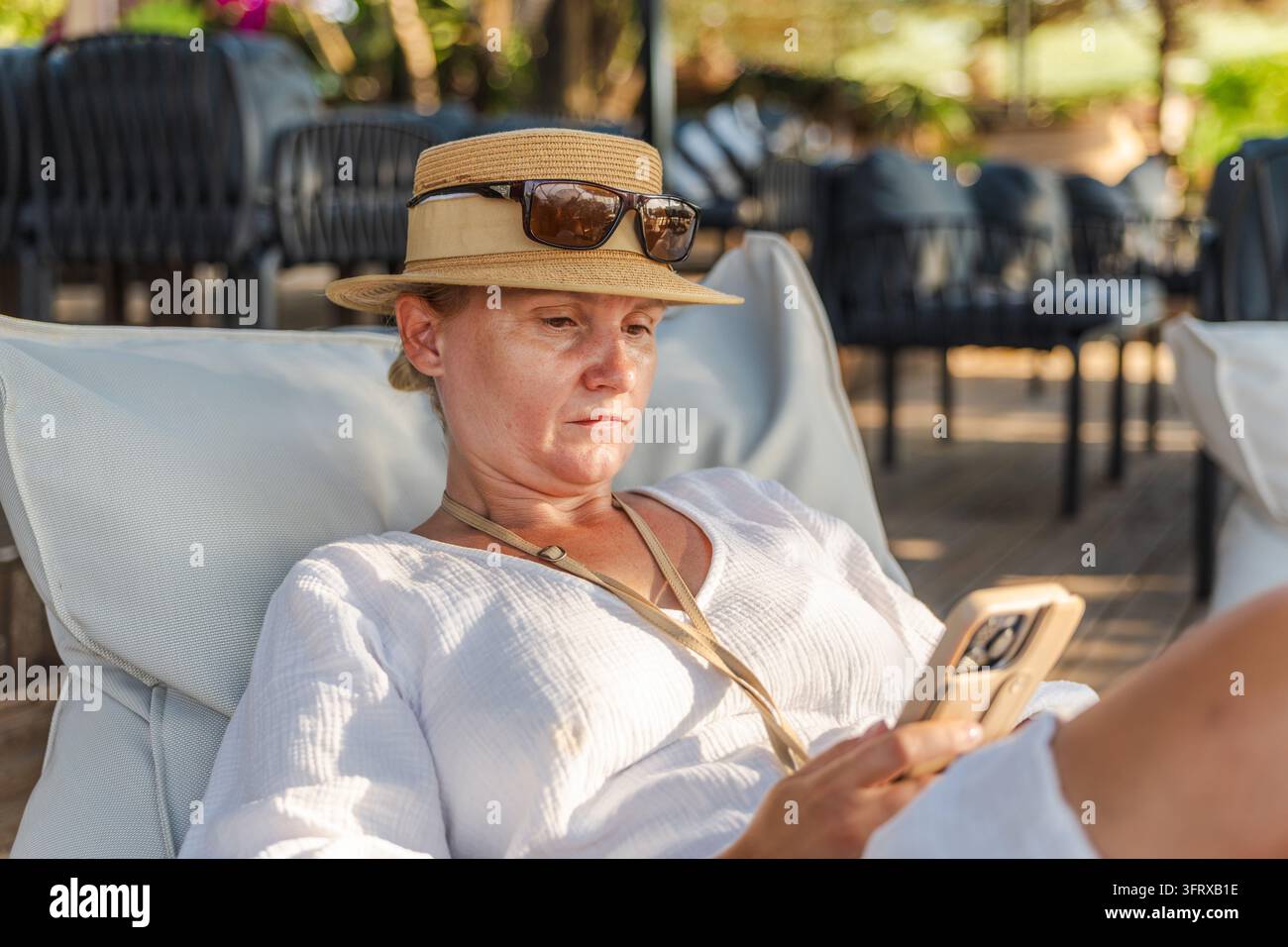Donna con cappello da sole si rilassa su una sedia da esterno mentre utilizza il suo smartphone. Foto Stock