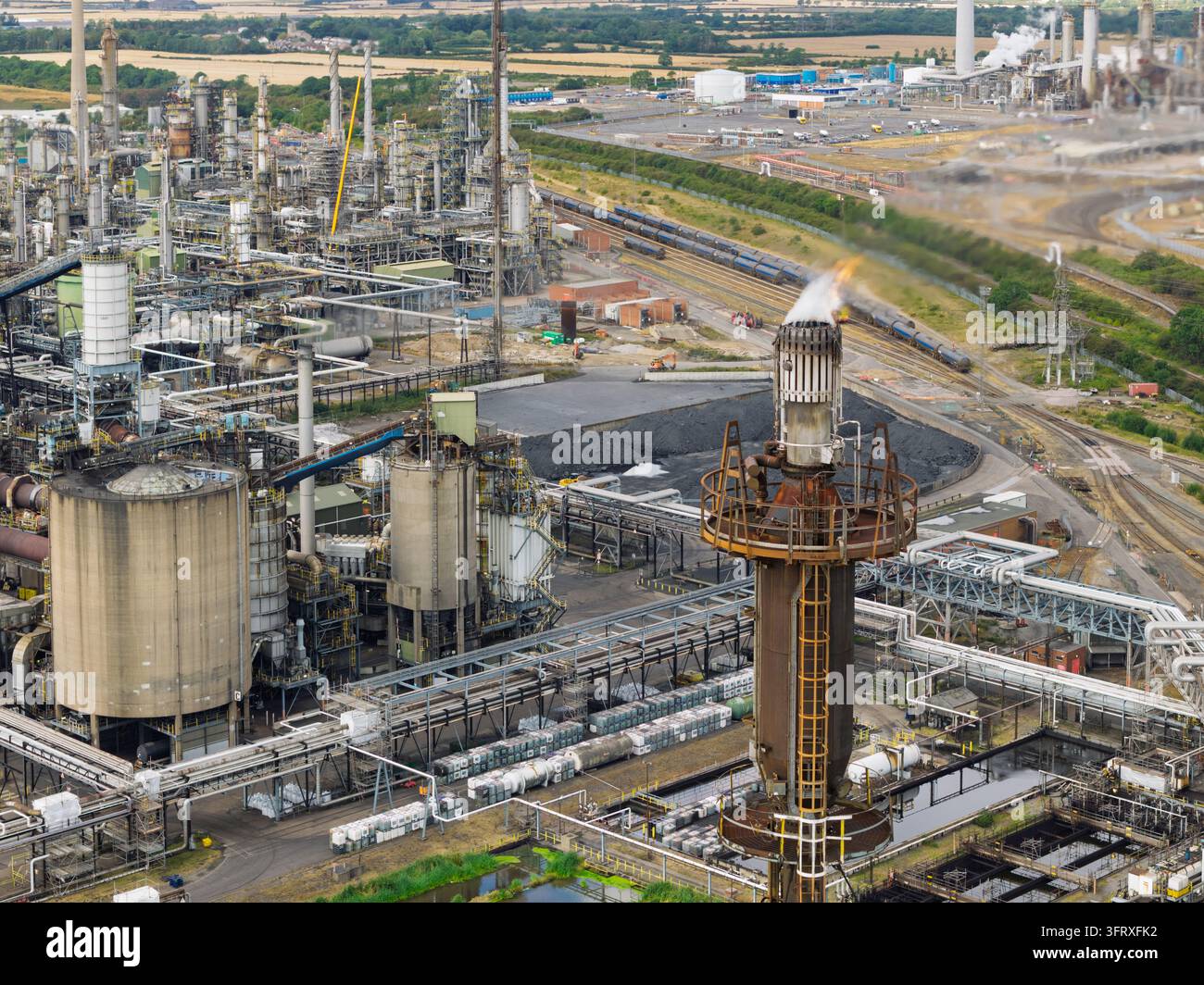 Infrastruttura industriale complessa di raffineria di petrolio con un alto camino di fiamma che brucia gas in eccesso sullo sfondo di binari ferroviari e serbatoi di stoccaggio su un Foto Stock