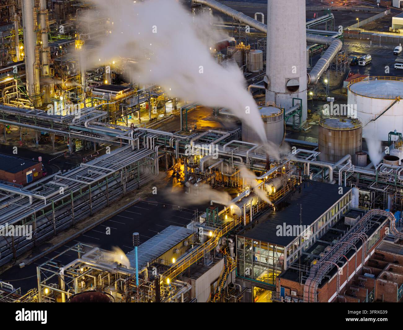 Grande complesso infrastrutturale di raffineria emette un pesante vapore bianco mentre opera a piena capacità sotto luci artificiali brillanti durante il crepuscolo Foto Stock